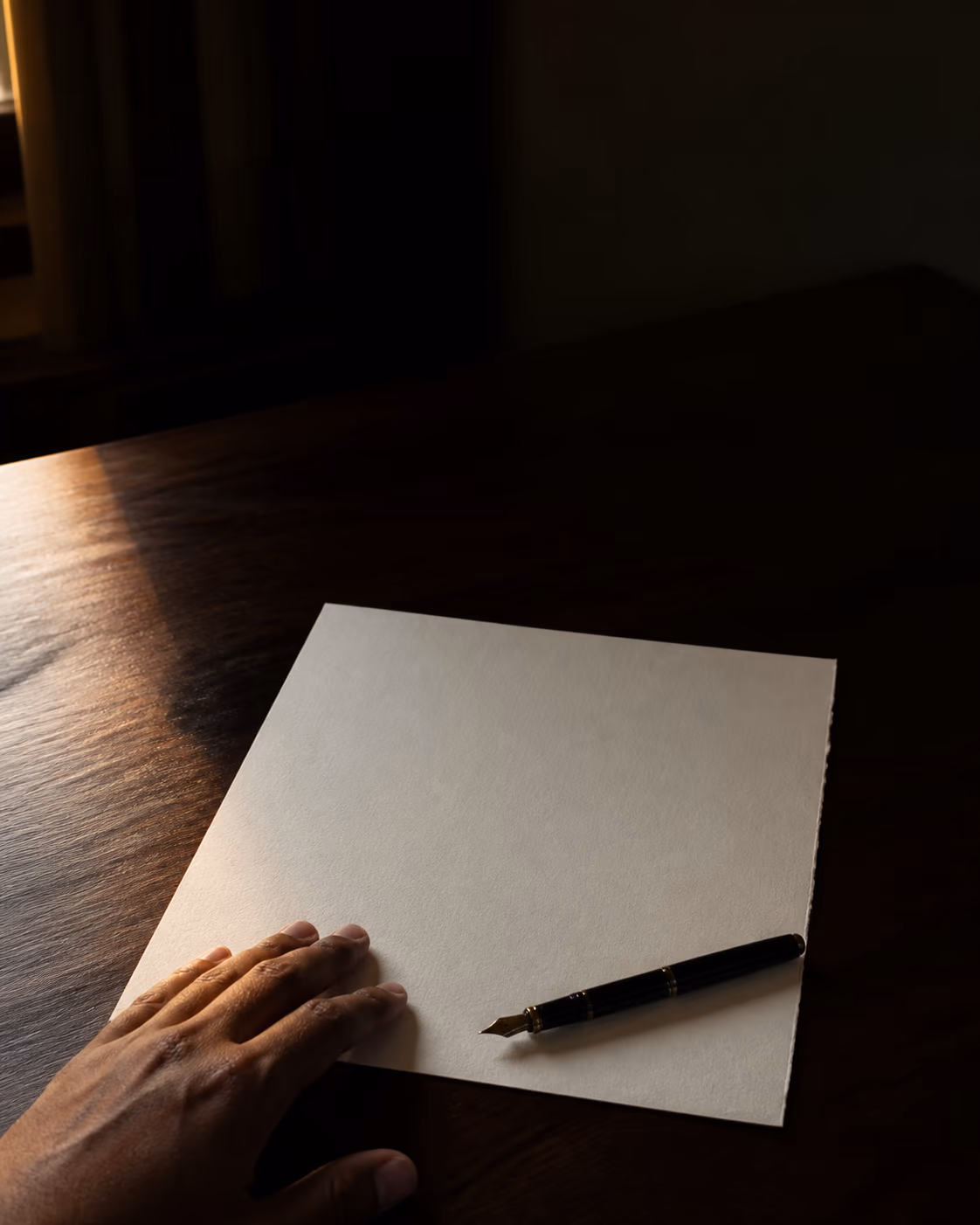 A single blank sheet of heavy cotton paper and a fountain pen on a dark table, lit by one low window.